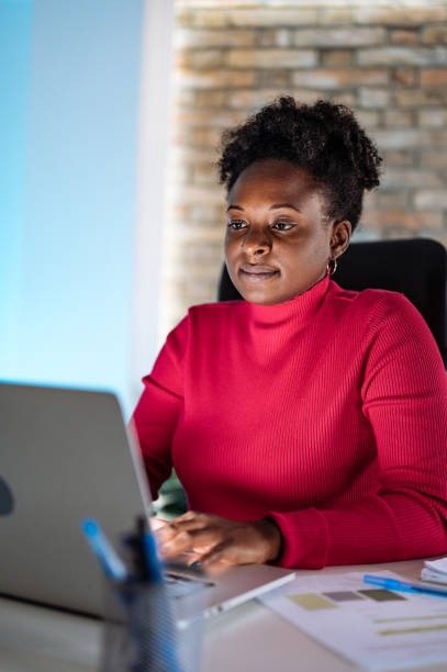 Woman in a red sweater working on her laptop at a desk with papers and blue pens, in a modern TechRay360 office with a brick wall background.